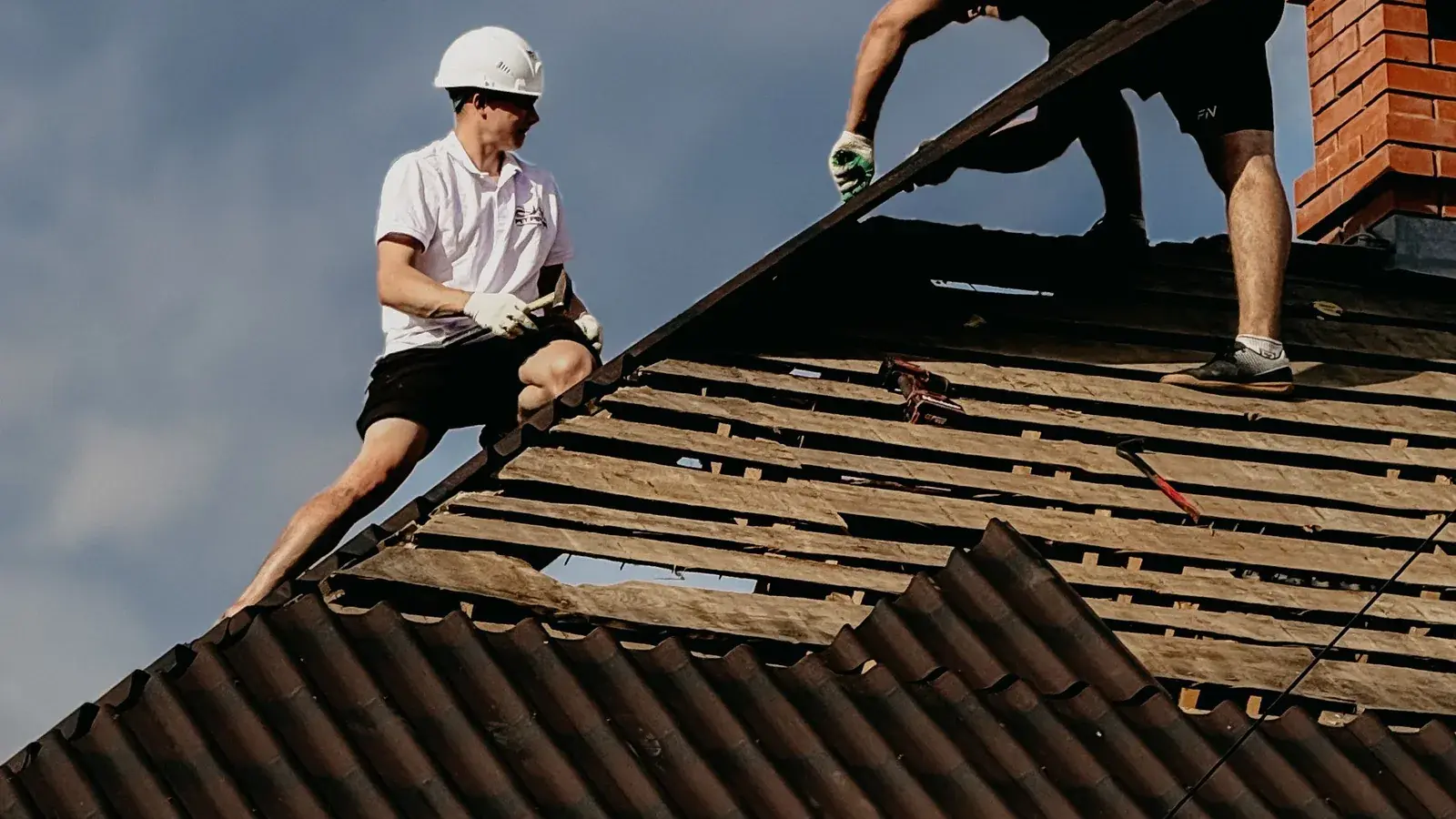 Two roofers stripping the old roof covering from a roof beside a brick chimney, exposing the battens beneath