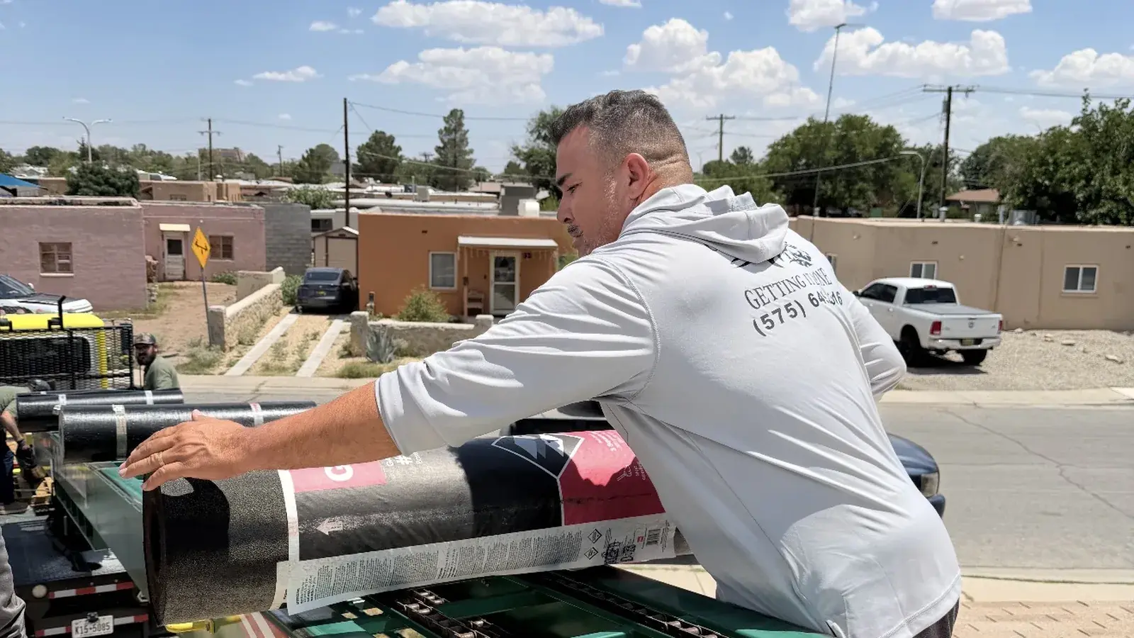 Gilbert & Sons crew member unloading rolls of roofing underlayment from a trailer on a Las Cruces street before a replacement job