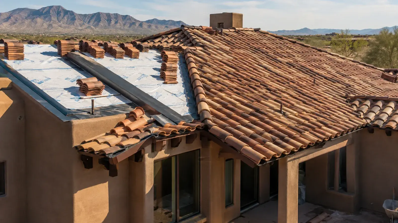 Clay barrel tile roof mid-installation on a Southwest home, with white underlayment exposed and tiles stacked in rows ready to lay