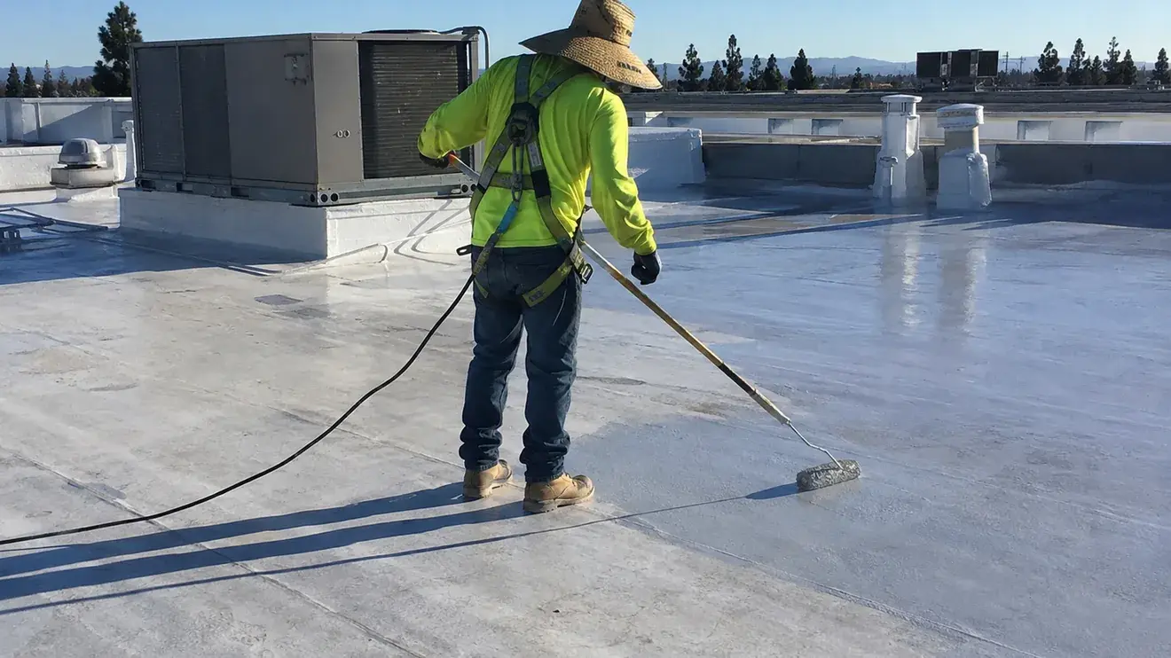 Roofer rolling a reflective coating across a commercial flat roof with rooftop HVAC units and a parapet behind