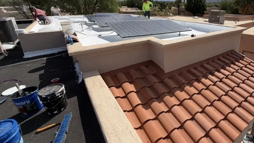 Coating buckets and a brush on a flat roof beside a freshly coated white surface, with crew working behind the parapet