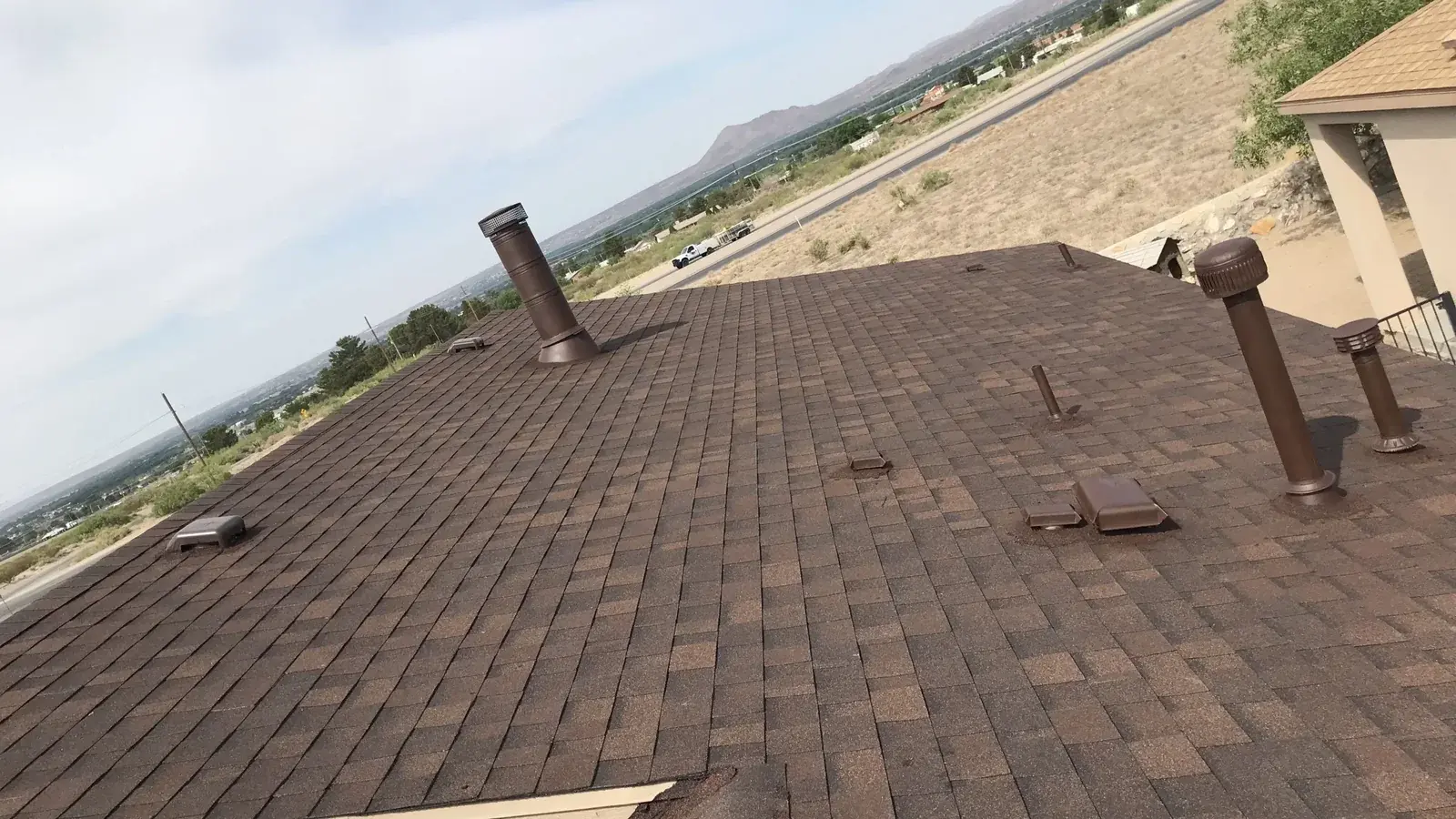 Brown asphalt shingle roof with several vent pipes and box vents, overlooking desert terrain