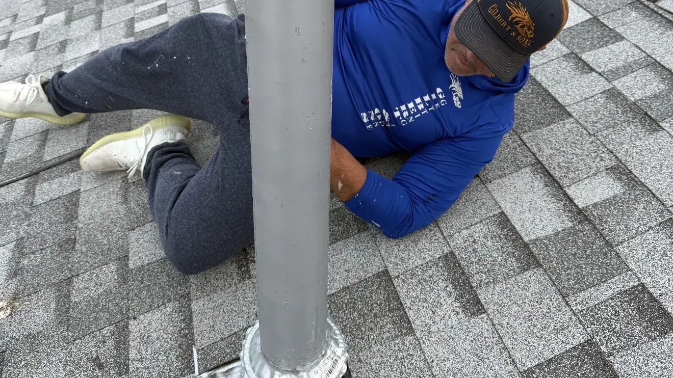 Roofer kneeling on an asphalt shingle roof to reseal the metal flashing boot around a vent pipe