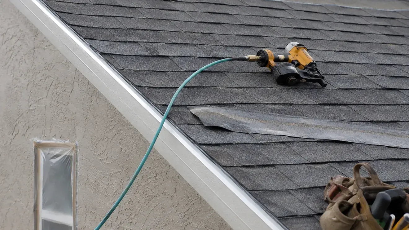 Nail gun resting on a shingled roof slope beside the drip edge, with a stucco wall alongside