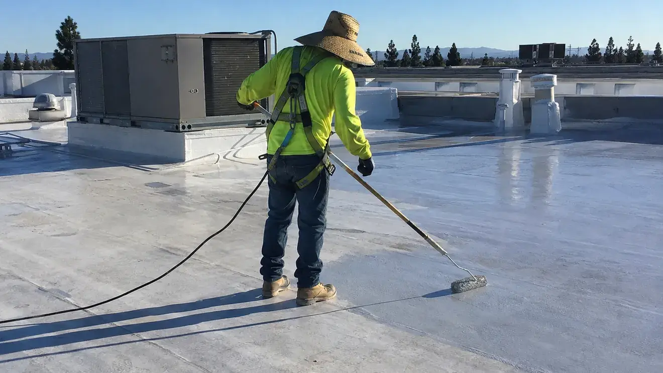 Roofer in a safety harness rolling silicone coating onto a commercial flat roof beside rooftop HVAC units