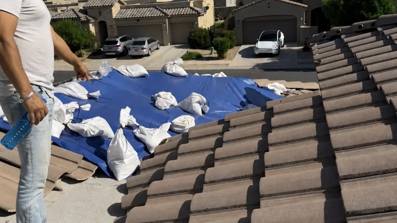 Concrete tile roof with a sandbagged emergency tarp over a damaged section, in a stucco-home neighbourhood