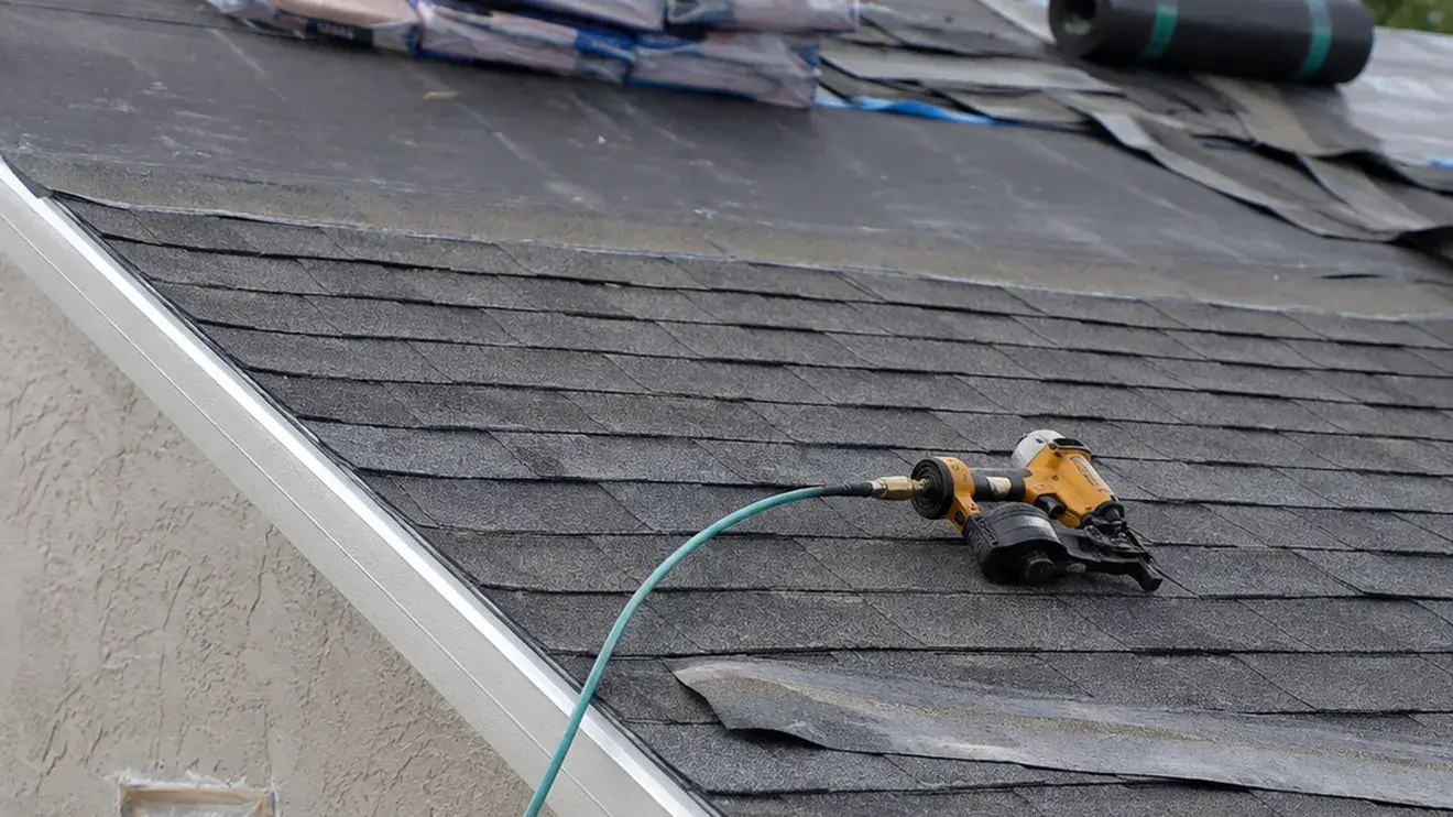 Nail gun resting on a partly shingled roof slope with drip edge, starter course, underlayment roll and shingle bundles staged at the ridge