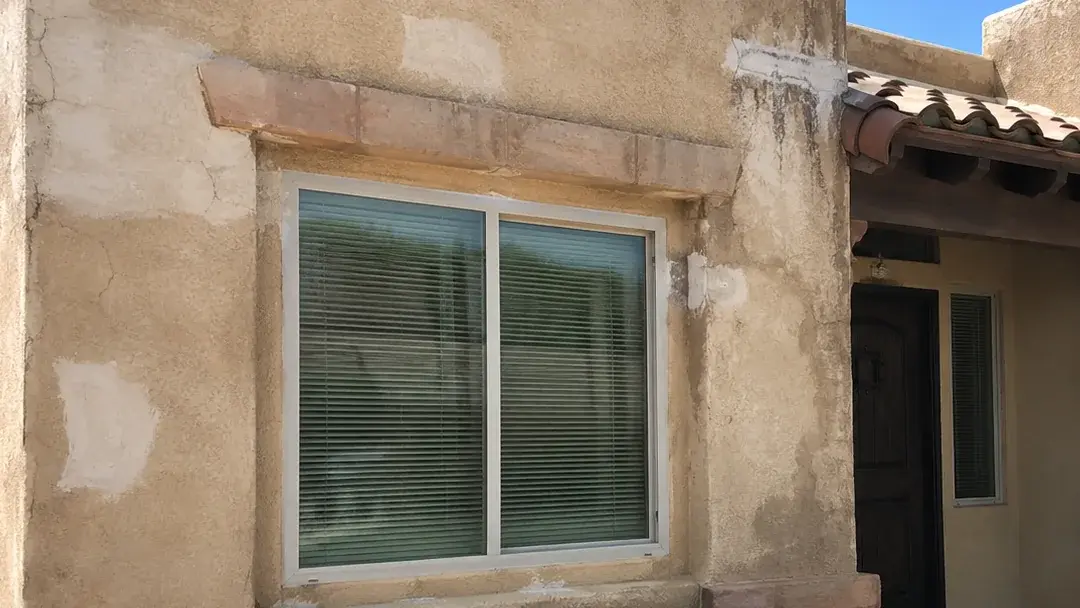 Close view of a stucco wall with fine cracking and mismatched patch repairs beside a window opening