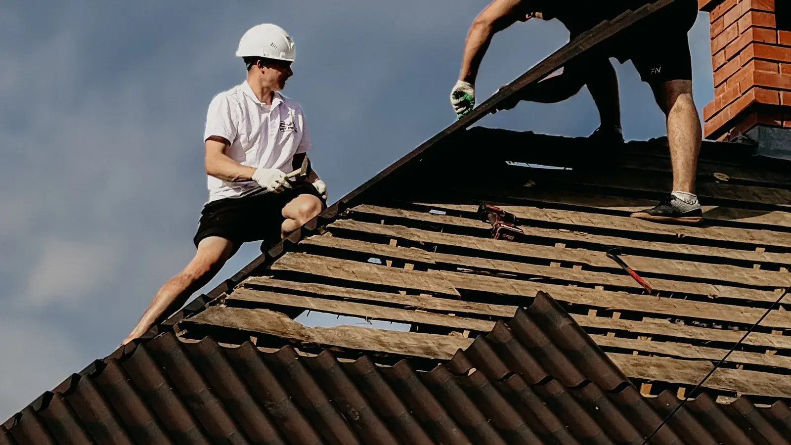 Two roofers stripping tiles from a roof slope, exposing the timber battens and deck underneath
