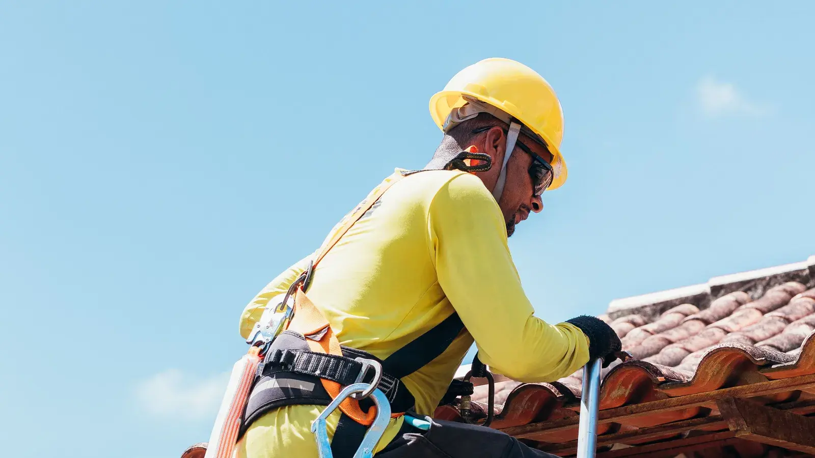 Roofer in a hard hat and fall-protection harness climbing from a ladder onto a clay tile roof above a stucco wall