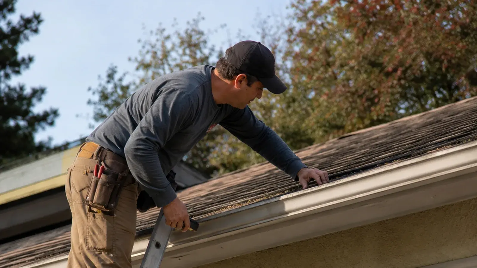 Roofer on a ladder checking the gutter and roof edge of a home during a scheduled maintenance visit