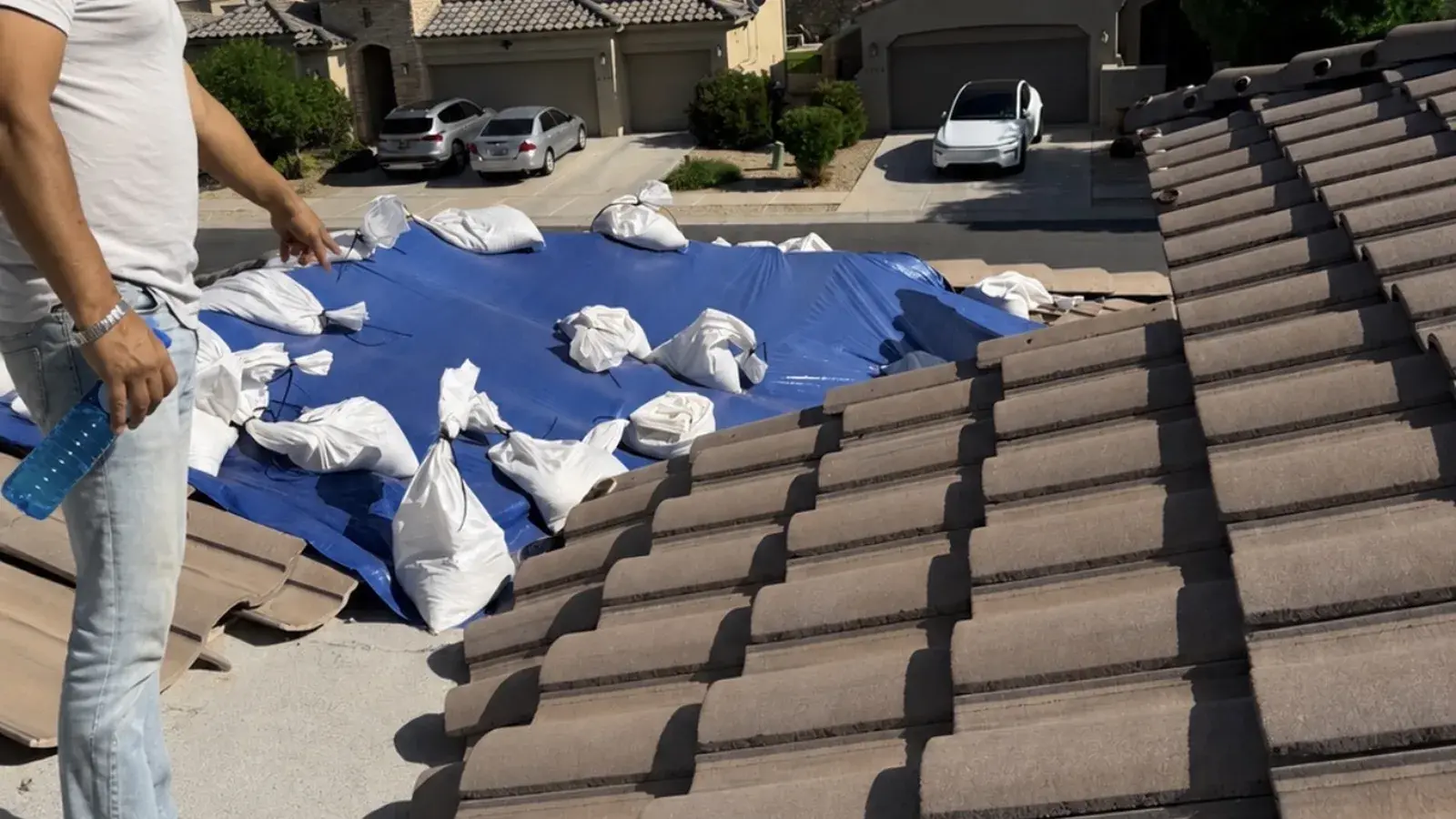 Roofer standing beside a sandbagged emergency tarp covering storm damage on a concrete tile roof in a Las Cruces neighbourhood