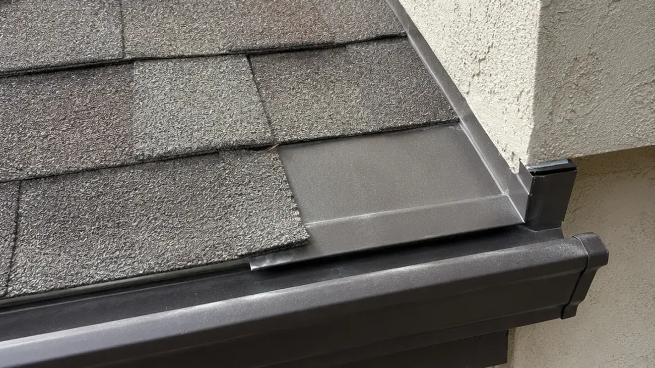 Close-up of metal flashing where an asphalt shingle roof meets a stucco wall, finished above a dark bronze gutter