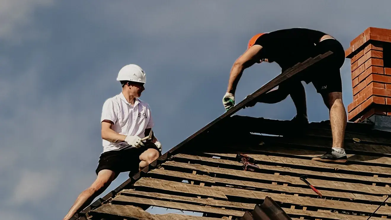 Two roofers removing roof covering beside a brick chimney, exposing the battens and deck underneath